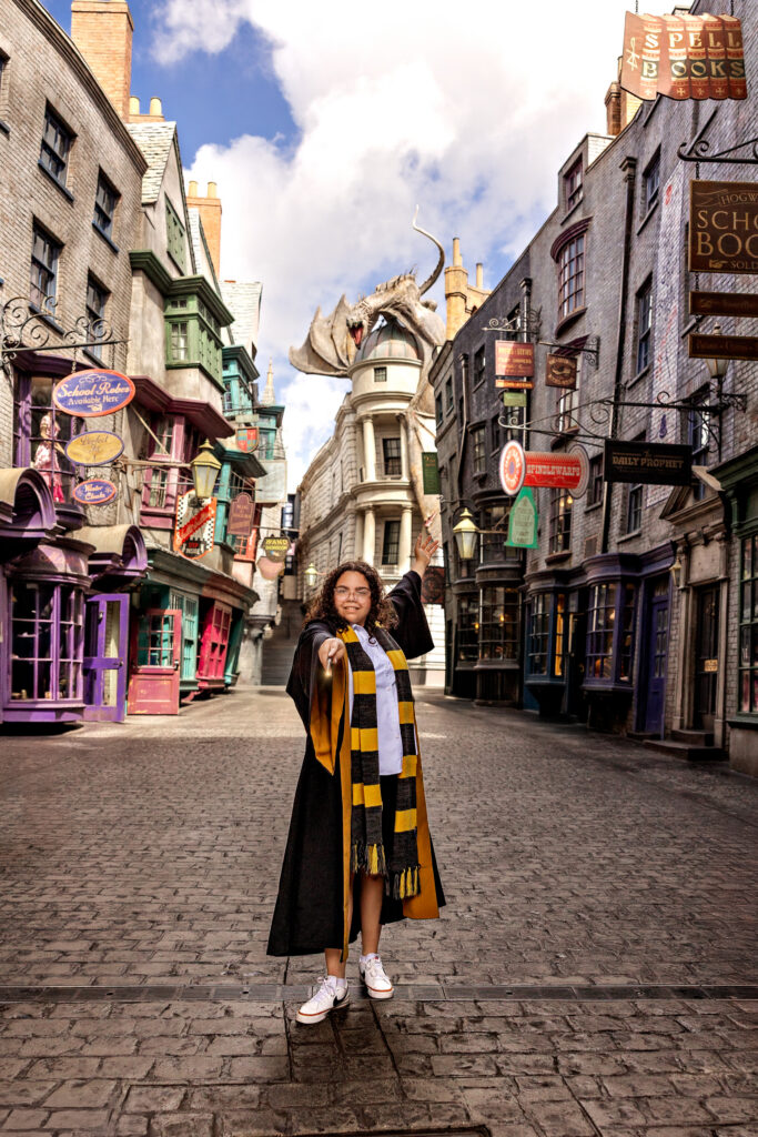 Haleigh in Hogwarts robes posing with her wand in Diagon Alley during her senior session at Universal Orlando, with the dragon and magical shops in the background