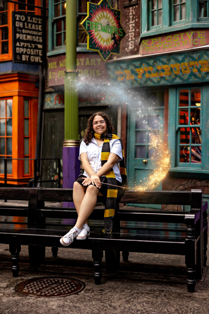 Haleigh smiling on a bench in Diagon Alley at Universal Orlando during her senior session, surrounded by magical golden sparkles and vibrant Harry Potter shop fronts