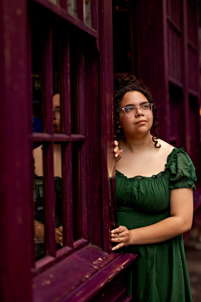 A medium close-up portrait of Haleigh looking thoughtfully to the side while standing in a purple doorway/window. She wears a flowing emerald green off-the-shoulder dress with ruffled sleeves, clear glasses, and has her curly hair styled in an updo. Her hands rest on the weathered purple door frame. Warm lighting and soft reflections enhance the moody, cinematic atmosphere of the Wizarding World setting.