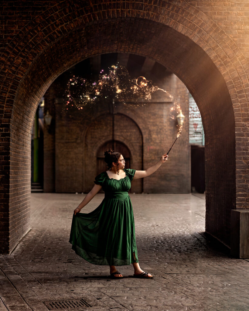 Haleigh in a green dress casting a magical spell with her wand under a brick archway at Universal Orlando during her senior photos, with sparkling effects.